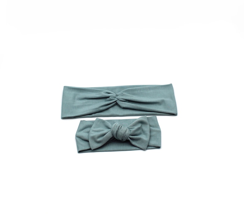 May include: Two teal headbands on a white background. One headband is a wide, twisted style. The other is a smaller headband with a bow. Both are made of a soft, stretchy fabric.