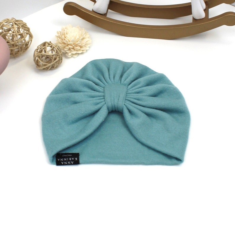 May include: A teal-colored baby turban hat with a large bow detail. The hat is made of soft fabric and features a small black label with white text that reads "ANNA KARINNA". The hat is displayed on a white surface with decorative items in the background.