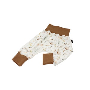 May include: Baby trousers featuring a white fabric adorned with a pattern of brown and green leaves. The cuffs and waistband are a solid brown. The trousers have a relaxed fit and a ruffled edge at the cuffs and waistband.