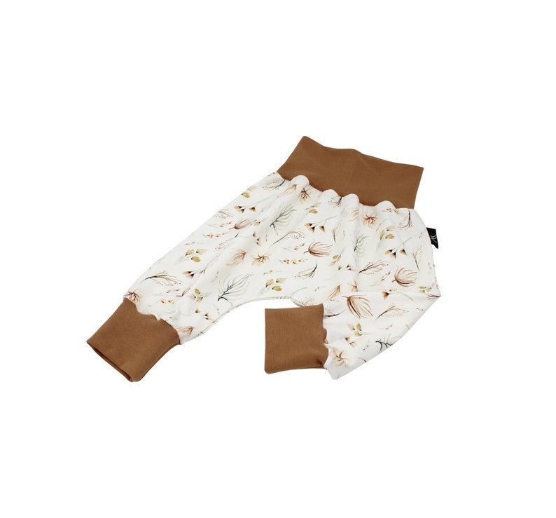 May include: Baby trousers featuring a white fabric adorned with a pattern of brown and green leaves. The cuffs and waistband are a solid brown. The trousers have a relaxed fit and a ruffled edge at the cuffs and waistband.