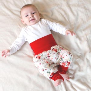 May include: A baby wearing a white long-sleeved top with a red waistband and floral print trousers. The trousers have red cuffs. The baby is lying on a white sheet.