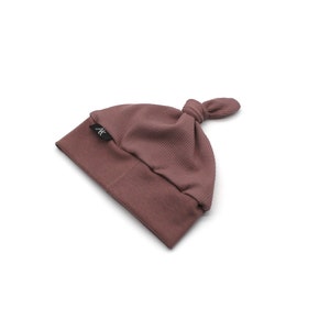 May include: A mauve-colored baby beanie with a knotted top. The hat is made of a ribbed knit fabric and features a small black label with white text. The hat is designed for infants and toddlers.