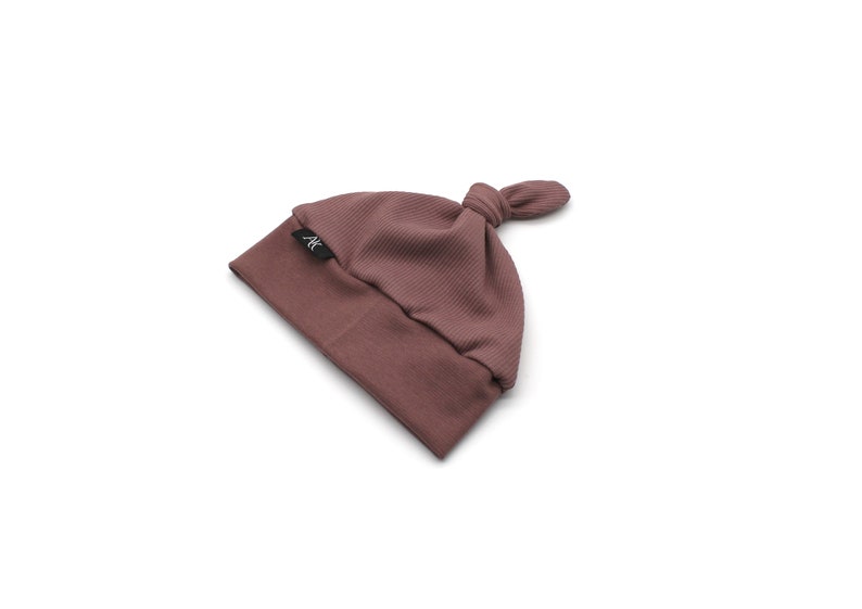 May include: A mauve-colored baby beanie with a knotted top. The hat is made of a ribbed knit fabric and features a small black label with white text. The hat is designed for infants and toddlers.