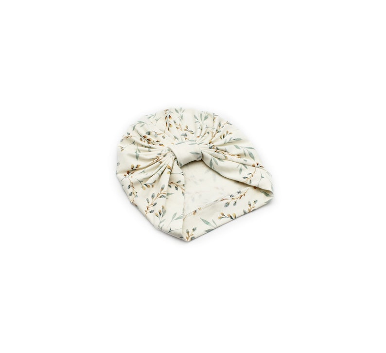 May include: A cream-colored baby turban with a botanical print of green and brown leaves and stems. The turban is knotted at the top and is made of soft fabric. The head covering is designed for infants.