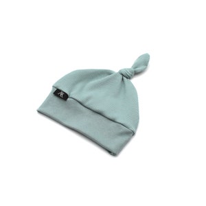May include: A light teal baby knot hat with a ribbed top and a smooth bottom. The hat has a small black tag with the letters "AK" in white. The hat is made of soft material.
