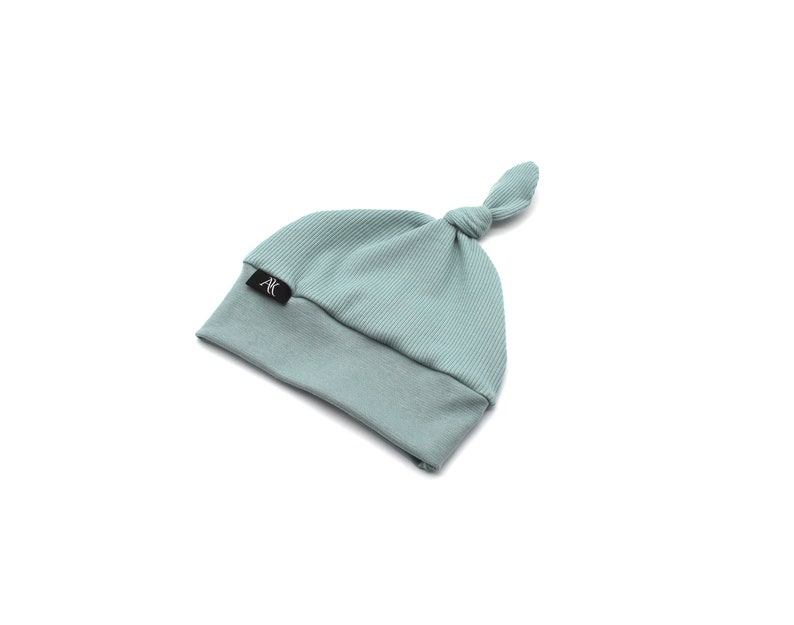 May include: A light teal baby knot hat with a ribbed top and a smooth bottom. The hat has a small black tag with the letters "AK" in white. The hat is made of soft material.