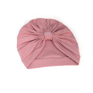 May include: A dusty rose turban-style hat. The hat is made of soft fabric and features a knotted detail at the top center. The head covering is designed to be worn on the head.