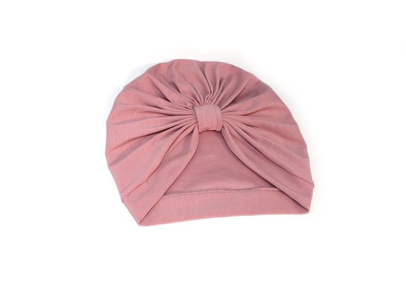 May include: A dusty rose turban-style hat. The hat is made of soft fabric and features a knotted detail at the top center. The head covering is designed to be worn on the head.