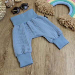 May include: Light blue baby harem pants with a gathered waistband and cuffs. The pants are made of a textured fabric and are designed for comfort. The pants are shown on a wooden surface with other baby accessories.