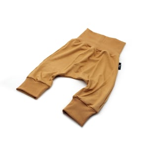 May include: Mustard-coloured baby trousers with a high waistband and ribbed cuffs. The soft, stretchy fabric is gathered at the crotch, creating a relaxed fit. A small black tag is sewn on the side.