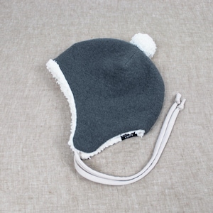 Peut inclure: Un bonnet en polaire gris fonc&eacute; avec une doublure et une bordure en sherpa blanc. Le bonnet pr&eacute;sente un pompon blanc sur le dessus, des oreillettes et deux longs liens en tissu clair. Une petite &eacute;tiquette noire avec du texte blanc est visible.