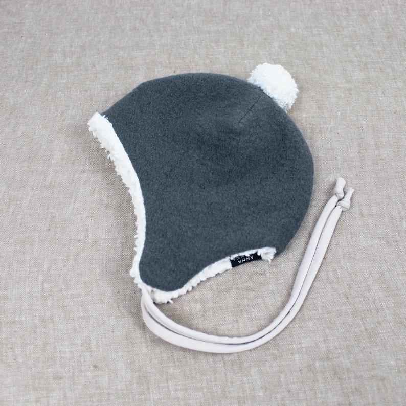 Peut inclure: Un bonnet en polaire gris fonc&eacute; avec une doublure et une bordure en sherpa blanc. Le bonnet pr&eacute;sente un pompon blanc sur le dessus, des oreillettes et deux longs liens en tissu clair. Une petite &eacute;tiquette noire avec du texte blanc est visible.