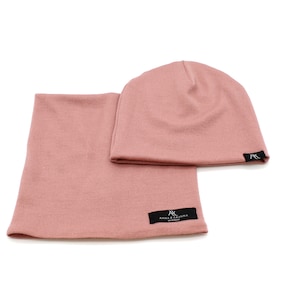 Może przedstawiać: Pasujący zestaw czapki i komina w kolorze pudrowego różu. Czapka ma prosty, dopasowany krój. Komin ma kształt tuby. Oba elementy mają małą czarną metkę z białym tekstem. Zestaw wykonany jest z miękkiej dzianiny.