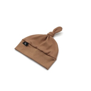 May include: A brown ribbed knit baby hat with a knotted top. The hat has a small black label with a white logo. The hat is made of a soft material and is designed for newborns.