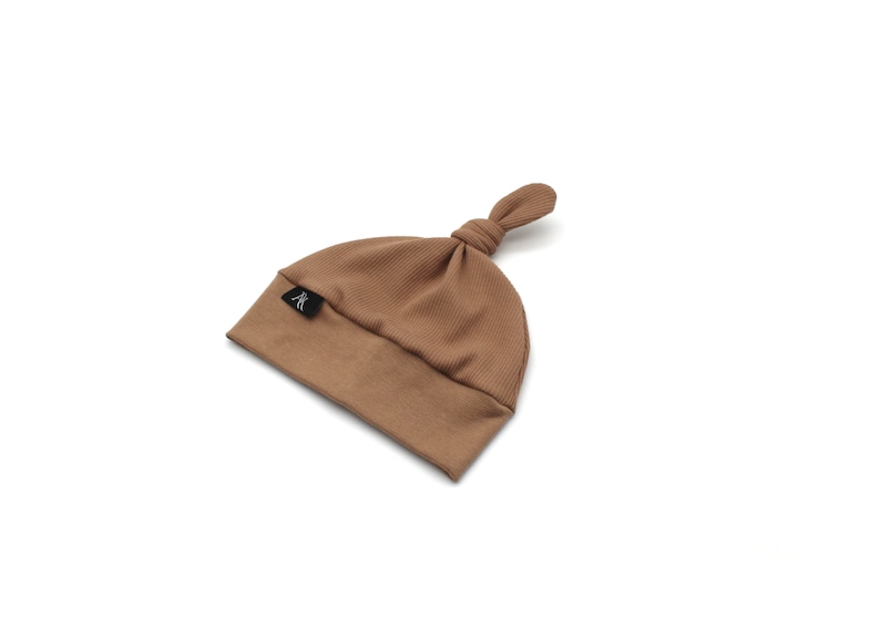 May include: A brown ribbed knit baby hat with a knotted top. The hat has a small black label with a white logo. The hat is made of a soft material and is designed for newborns.