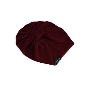 May include: A burgundy turban-style hat with a knotted detail at the crown. The hat is made of a ribbed fabric and features a small black tag with white text. The hat is designed to be worn on the head.