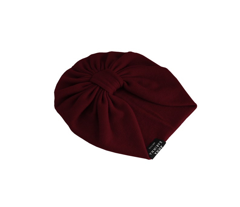 May include: A burgundy turban-style hat with a knotted detail at the crown. The hat is made of a ribbed fabric and features a small black tag with white text. The hat is designed to be worn on the head.