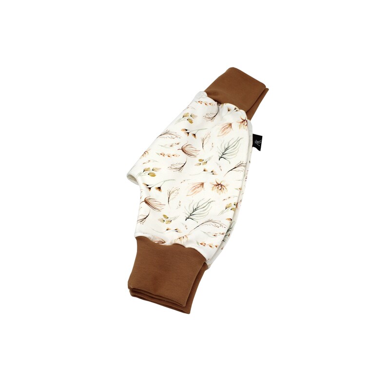 May include: Baby trousers with a cream-coloured background and a pattern of brown and green leaves and feathers. The cuffs are a solid brown colour. The trousers are made of soft fabric.