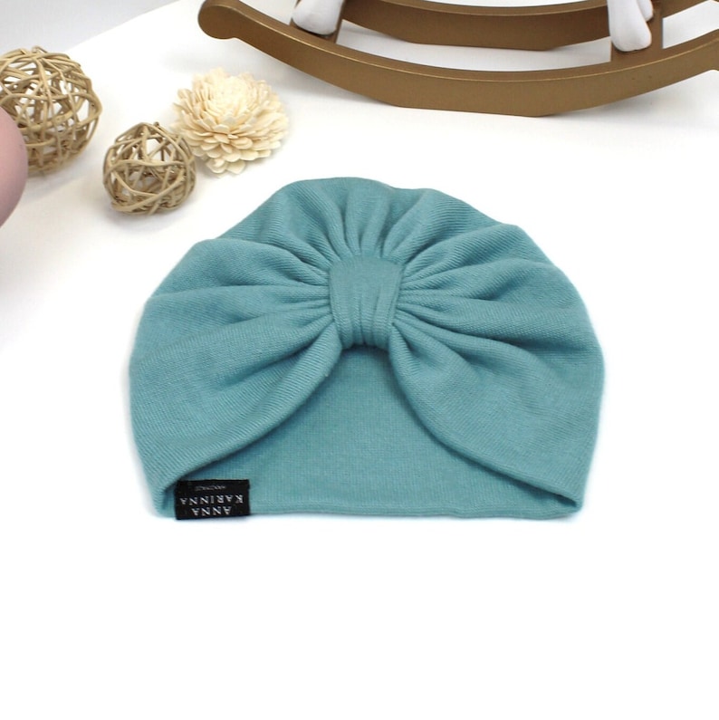 Może przedstawiać: Turkusowy turban dla dziecka z dużym detalem w postaci kokardy. Kapelusz wykonany jest z miękkiej tkaniny i posiada małą czarną metkę z napisem "ANNA KARINNA". Turban przeznaczony jest dla niemowląt i małych dzieci.