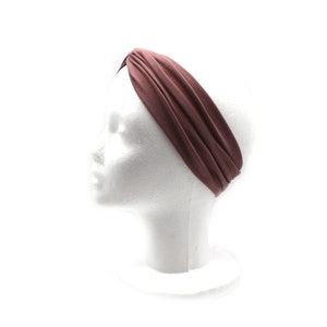 May include: A mauve-colored headband is displayed on a white mannequin head. The wide, twisted headband is made of a soft fabric. The background is plain white, highlighting the headband's color and design. This accessory is suitable for various hairstyles.