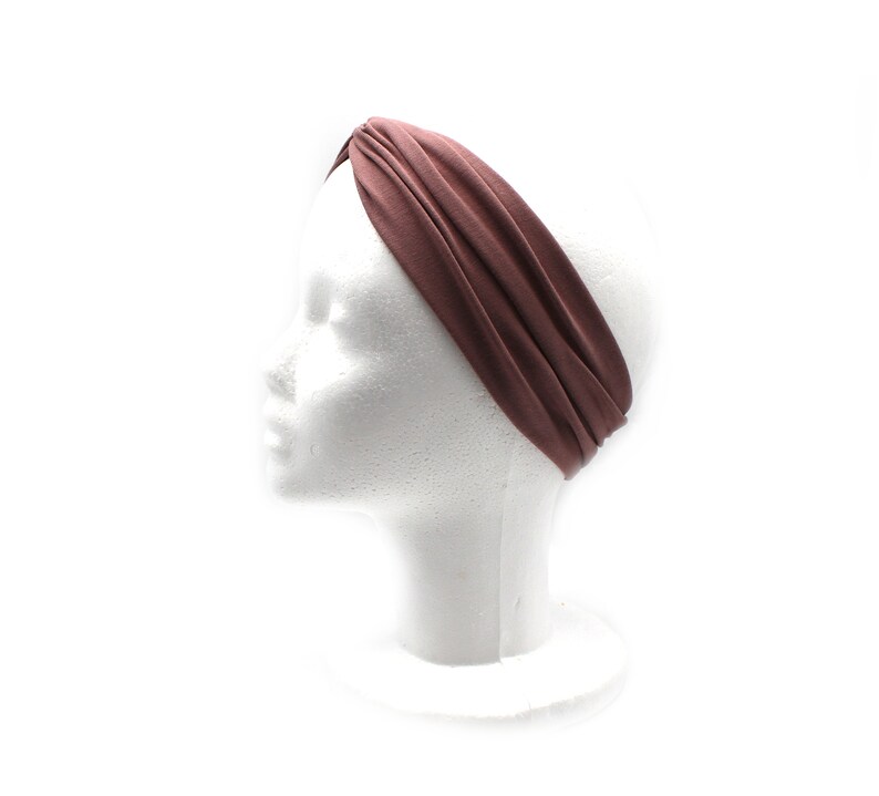May include: A mauve-colored headband is displayed on a white mannequin head. The wide, twisted headband is made of a soft fabric. The background is plain white, highlighting the headband's color and design. This accessory is suitable for various hairstyles.