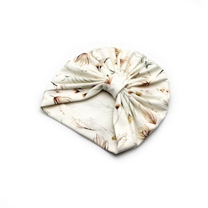 May include: Cream-colored turban with a floral pattern in shades of brown and green. The fabric is soft and gathered at the top, creating a stylish head covering. The turban is designed for comfort and fashion.