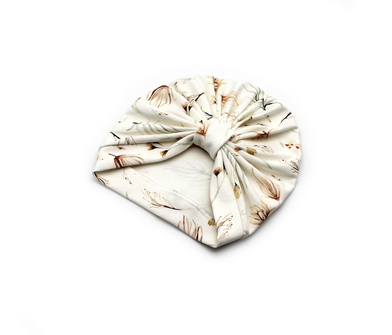 May include: Cream-colored turban with a floral pattern in shades of brown and green. The fabric is soft and gathered at the top, creating a stylish head covering. The turban is designed for comfort and fashion.