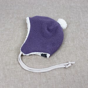 Peut inclure: Un bonnet violet fonc&eacute; &agrave; la texture granuleuse, dot&eacute; d'une doublure en polaire blanche sur les bords et d'un pompon blanc sur le dessus. Le bonnet comprend deux longs liens de couleur claire et une petite &eacute;tiquette noire avec le texte Anna Karinna.
