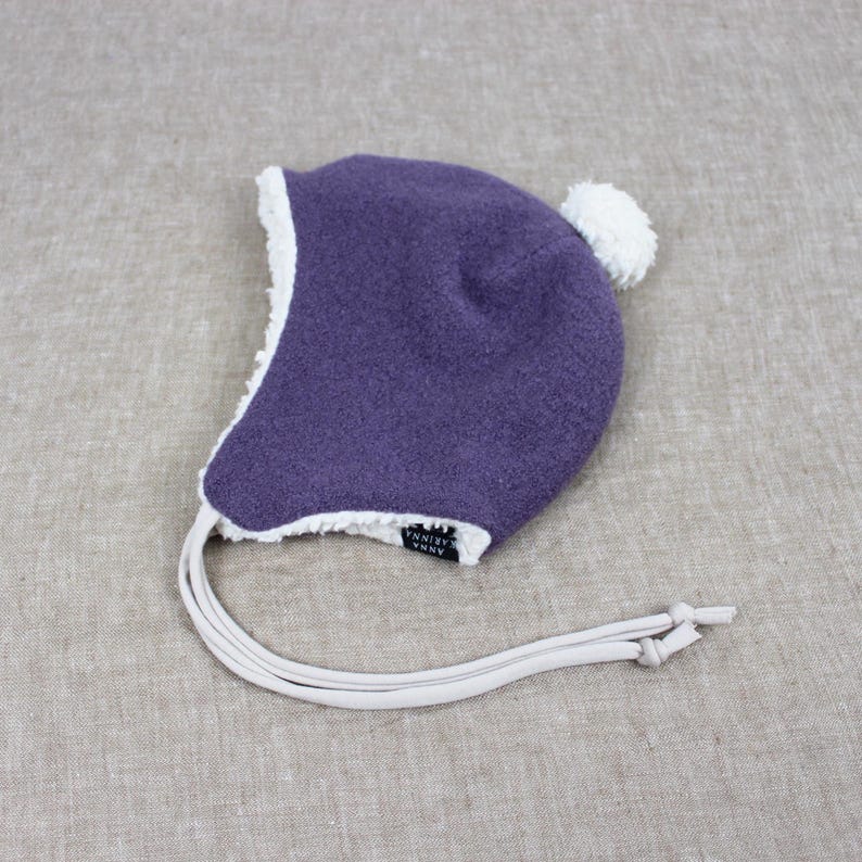 Peut inclure: Un bonnet violet fonc&eacute; &agrave; la texture granuleuse, dot&eacute; d'une doublure en polaire blanche sur les bords et d'un pompon blanc sur le dessus. Le bonnet comprend deux longs liens de couleur claire et une petite &eacute;tiquette noire avec le texte Anna Karinna.