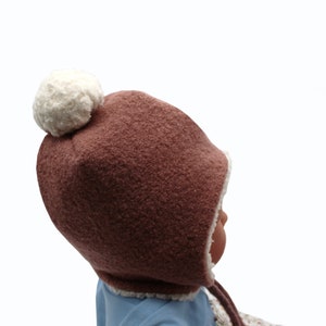 Peut inclure: Un bonnet marron textur&eacute; avec un pompon blanc sur le dessus et une doublure en polaire blanche. Le bonnet pr&eacute;sente des oreillettes et des liens marron qui pendent sur les c&ocirc;t&eacute;s. Le tissu ext&eacute;rieur a un aspect duveteux et la bordure blanche est visible.