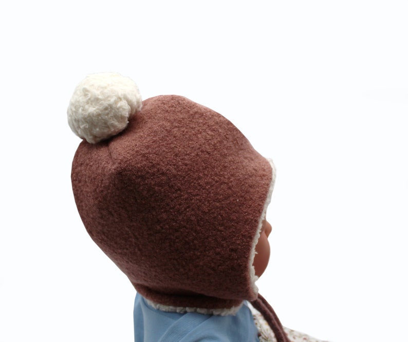 Peut inclure: Un bonnet marron textur&eacute; avec un pompon blanc sur le dessus et une doublure en polaire blanche. Le bonnet pr&eacute;sente des oreillettes et des liens marron qui pendent sur les c&ocirc;t&eacute;s. Le tissu ext&eacute;rieur a un aspect duveteux et la bordure blanche est visible.