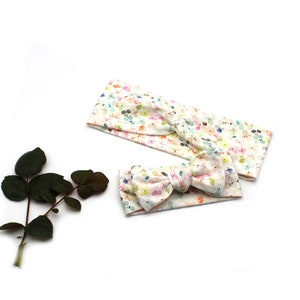 May include: Two headbands with a floral pattern. One is a wide headband, and the other is a smaller headband with a bow. The floral pattern includes pastel colors like pink, yellow, and blue on a white background. A dark green leafy stem is in the image.