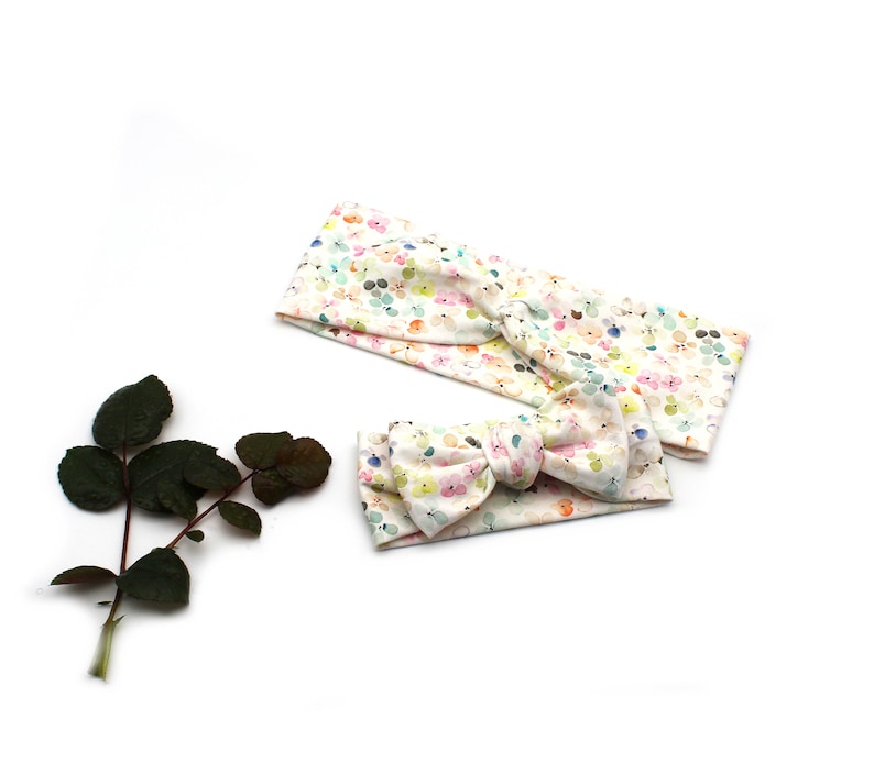 May include: Two headbands with a floral pattern. One is a wide headband, and the other is a smaller headband with a bow. The floral pattern includes pastel colors like pink, yellow, and blue on a white background. A dark green leafy stem is in the image.