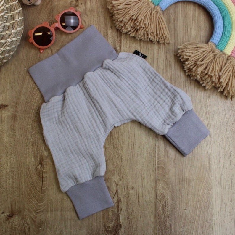 May include: Light gray baby pants with a wide waistband and ribbed cuffs. The pants are made of a soft, textured fabric. The pants are laying on a wooden surface, with a pair of pink sunglasses and a rainbow decoration in the background.