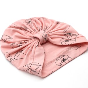 May include: A light pink turban-style hat with a floral pattern in black outlines. The hat is made of soft fabric and features a knotted design at the top. The floral design is repeated across the surface.