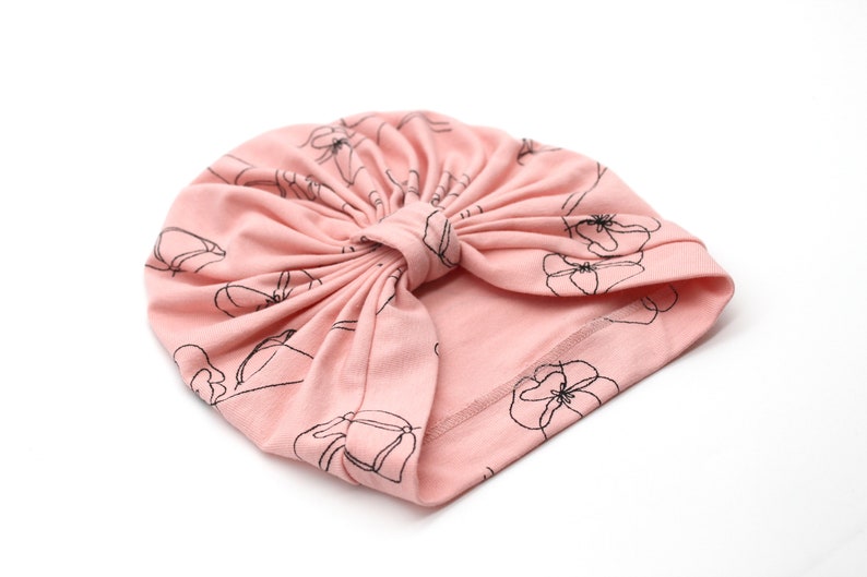 May include: A light pink turban-style hat with a floral pattern in black outlines. The hat is made of soft fabric and features a knotted design at the top. The floral design is repeated across the surface.