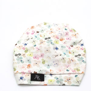 May include: A soft, floral-patterned beanie hat. The hat features a white background with a repeating pattern of pastel-colored flowers in pink, blue, green, and yellow. A small black tag with the letters "AK" is sewn on the brim.