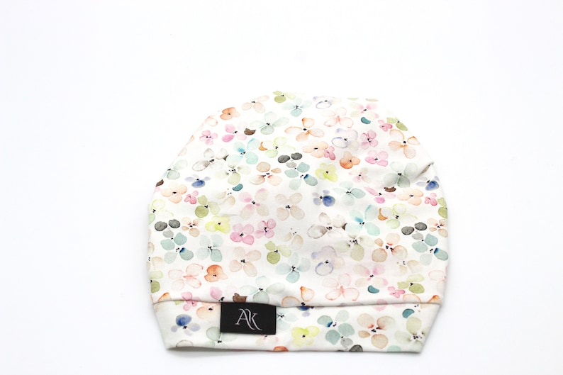 May include: A soft, floral-patterned beanie hat. The hat features a white background with a repeating pattern of pastel-colored flowers in pink, blue, green, and yellow. A small black tag with the letters "AK" is sewn on the brim.