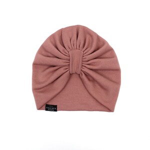 Może przedstawiać: R&oacute;żowy turban w kolorze pudrowego r&oacute;żu. Kapelusz wykonany jest z miękkiej, prążkowanej tkaniny i posiada marszczony detal w postaci węzła pośrodku. Mała czarna metka z białym tekstem jest wszyta z boku. Kapelusz jest przeznaczony do noszenia na głowie.