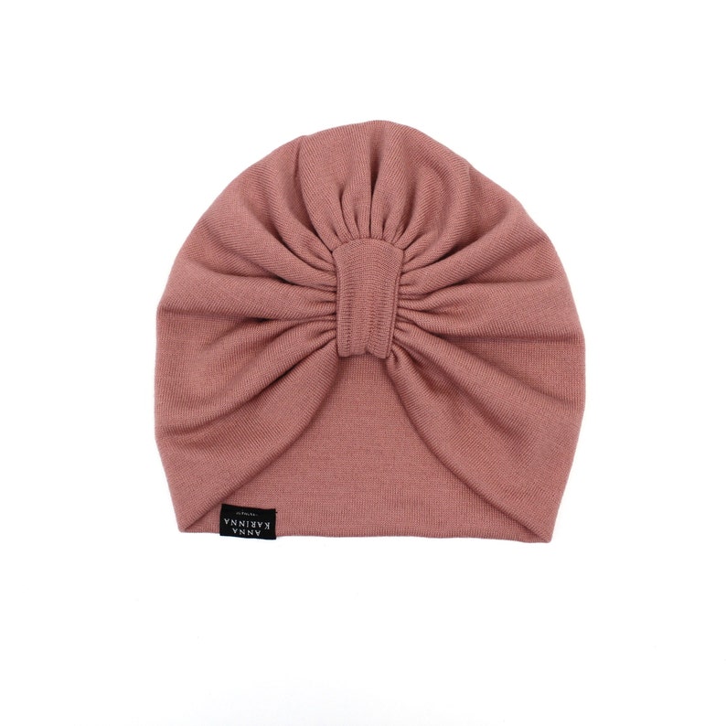 Może przedstawiać: R&oacute;żowy turban w kolorze pudrowego r&oacute;żu. Kapelusz wykonany jest z miękkiej, prążkowanej tkaniny i posiada marszczony detal w postaci węzła pośrodku. Mała czarna metka z białym tekstem jest wszyta z boku. Kapelusz jest przeznaczony do noszenia na głowie.