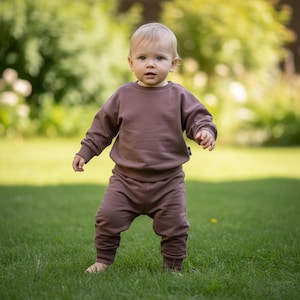Könnte beinhalten: Ein einfarbiges braunes Set bestehend aus einem langärmeligen Sweatshirt und einer passenden Jogginghose. Das Sweatshirt hat einen Rundhalsausschnitt und lange Ärmel mit gerippten Bündchen. Die Hose ist locker geschnitten und hat gerippte Beinabschlüsse.