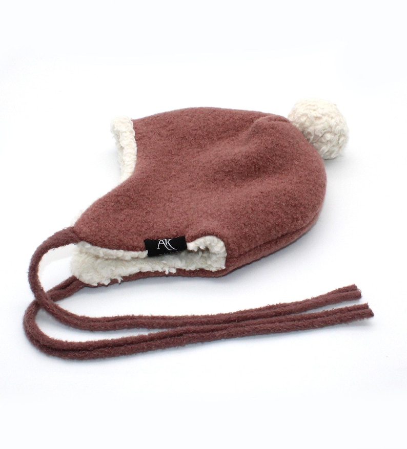 Peut inclure: Un bonnet &agrave; oreillettes marron avec une doublure en sherpa cr&egrave;me et un pompon cr&egrave;me assorti sur le dessus. Le bonnet pr&eacute;sente de longs liens marron et une petite &eacute;tiquette en tissu noir avec les lettres blanches AK cousues sur le c&ocirc;t&eacute;.