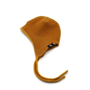 May include: A mustard yellow bonnet crafted from a textured, felted fabric. The bonnet features a rounded crown, ear flaps that extend into long chin ties, and a small black rectangular label with the white letters AK sewn on the side.
