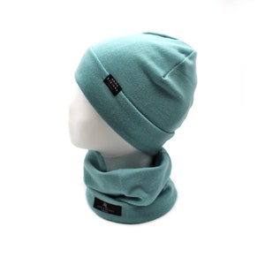 May include: A teal beanie hat and matching neck gaiter set. The hat and gaiter are made of a soft fabric and feature small black labels with white text. The set is displayed on a white mannequin head against a white background.