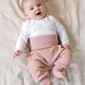 May include: A baby wearing a white long-sleeved top and pink ribbed trousers. The baby is lying on a light-coloured sheet and looking at the camera.