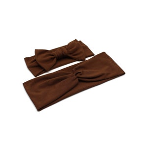 May include: Two brown headbands on a white background. One headband has a bow, and the other is a twist style. The headbands are made of a soft fabric and are a solid brown color.