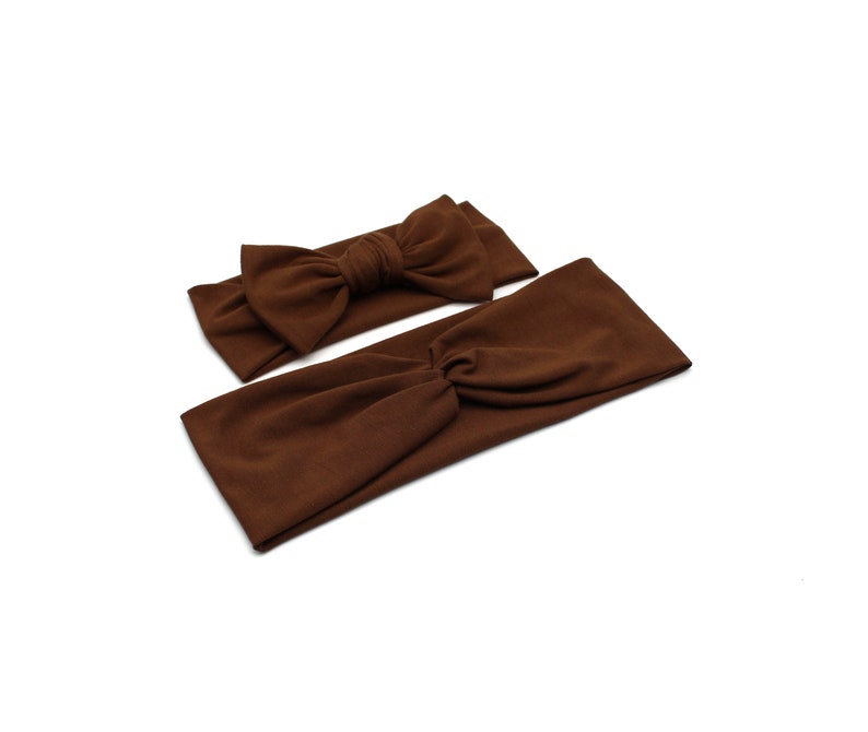 May include: Two brown headbands on a white background. One headband has a bow, and the other is a twist style. The headbands are made of a soft fabric and are a solid brown color.