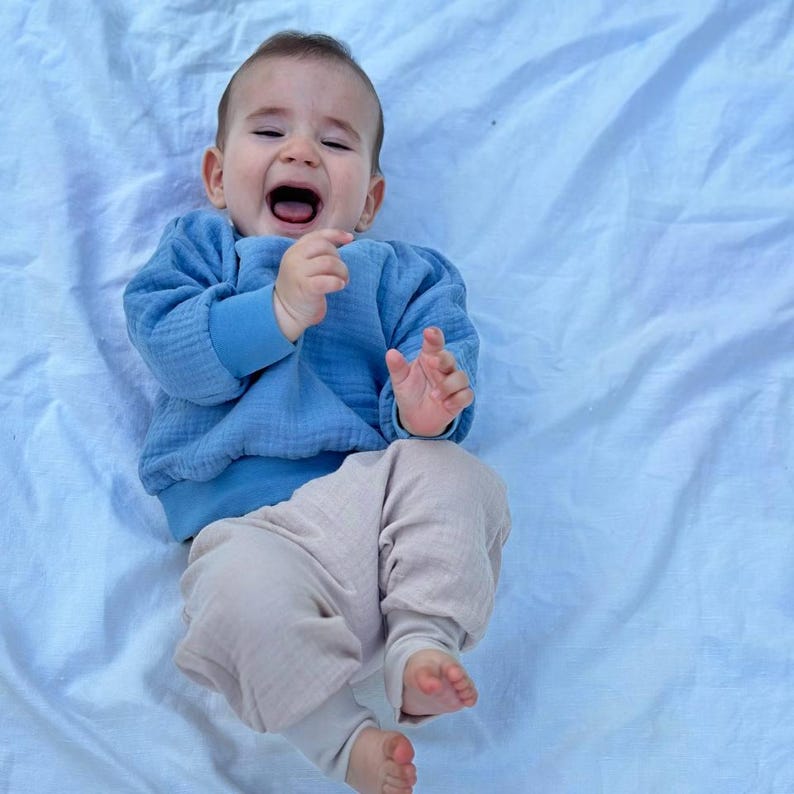 May include: A baby wearing a blue long-sleeved shirt and beige pants lies on a light blue blanket. The baby is crying with its mouth open and arms raised. The clothing appears to be made of a soft, textured fabric.
