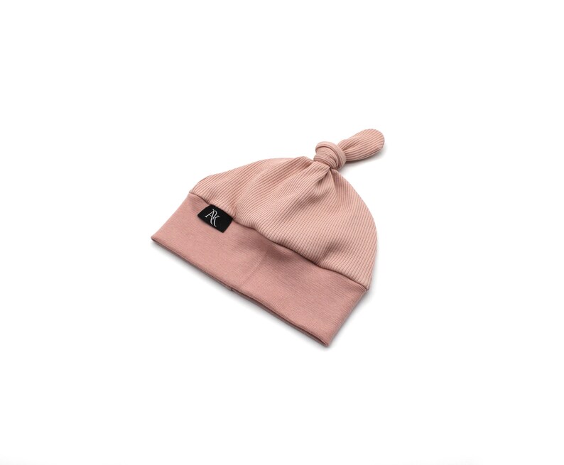 May include: A light pink baby beanie hat with a ribbed crown and a knotted top. The hat features a folded brim and a small black label with white text. The hat is made of a soft, stretchy material.
