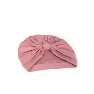 May include: A dusty rose turban-style hat. The hat is made of soft fabric and features a gathered design at the top, creating a stylish, knotted effect. The head covering is set against a white background.